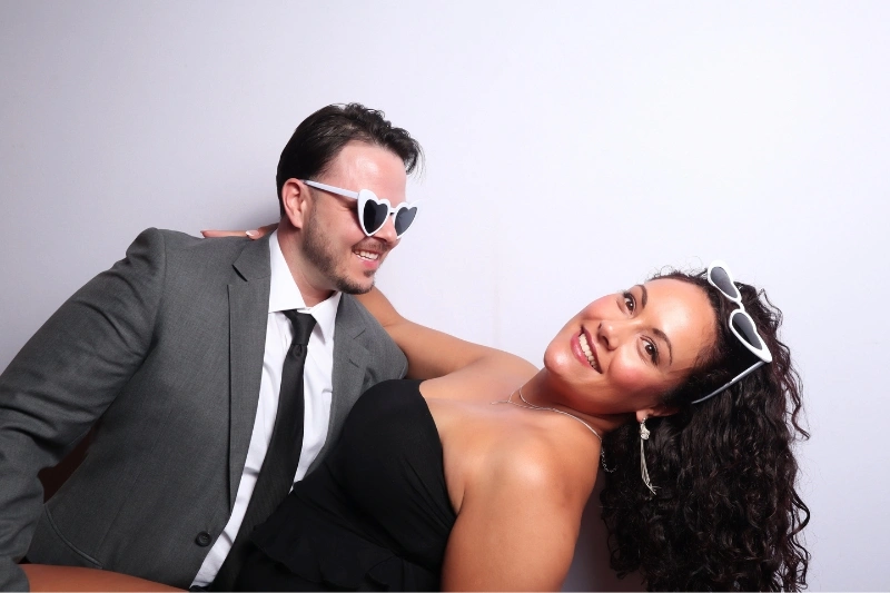 Smiling man in gray suit and heart-shaped sunglasses leaning towards a smiling woman in a black strapless dress and matching heart-shaped sunglasses.