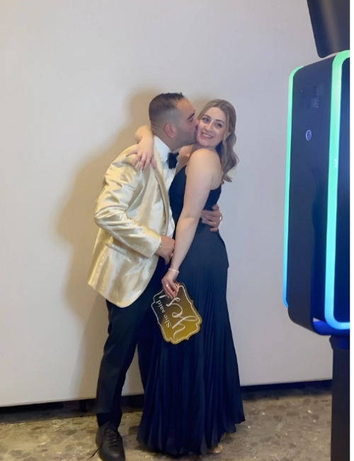 Man in gold blazer kissing a woman in a long black dress while she holds a decorative sign.