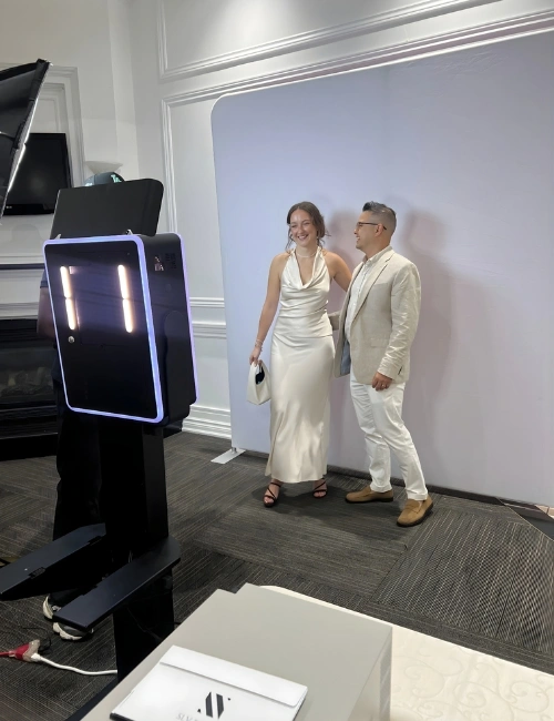 A woman in a white dress and a man in a light-colored suit pose together in front of a photo booth backdrop inside a room.