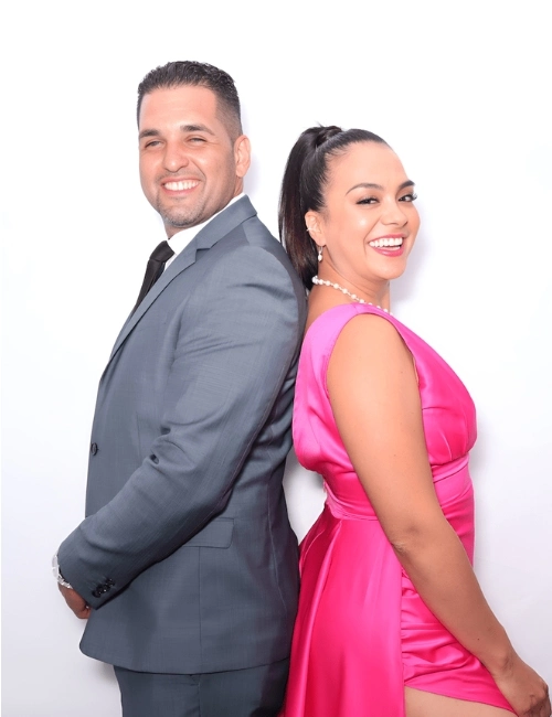 Smiling man in a gray suit and woman in a bright pink dress standing back to back against a white background.