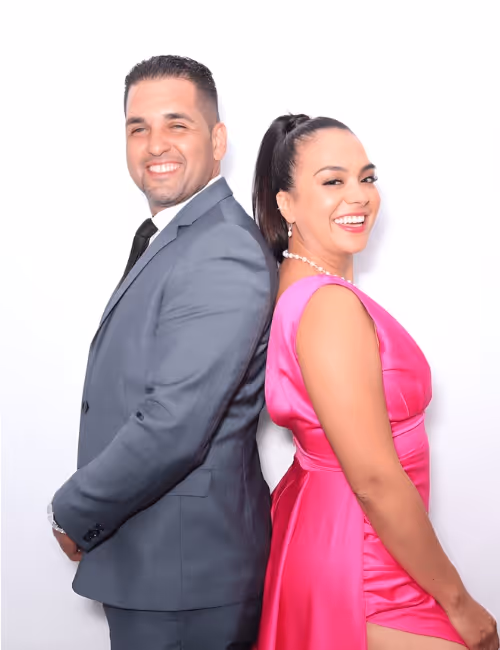 Smiling man in a gray suit and woman in a bright pink dress standing back to back against a white background.
