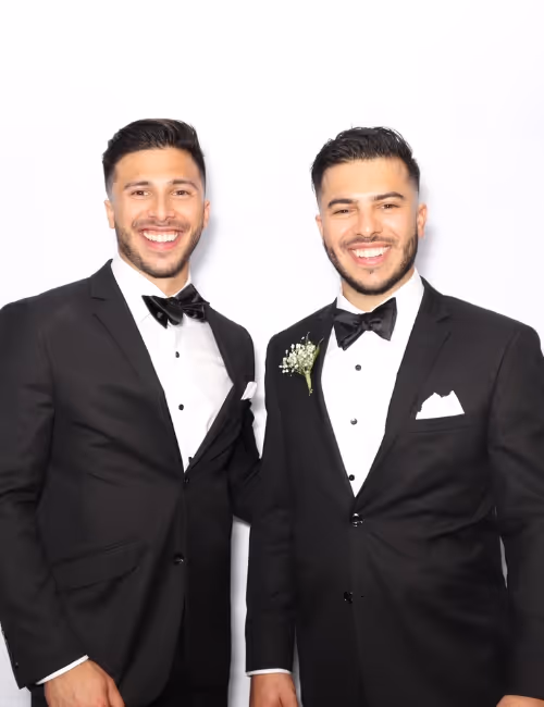 Two men wearing black tuxedos with bow ties smiling against a white background, one with a boutonniere.