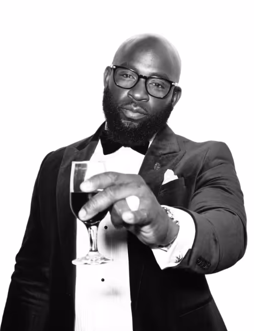 Man wearing glasses and a tuxedo, holding a glass of dark wine toward the camera.