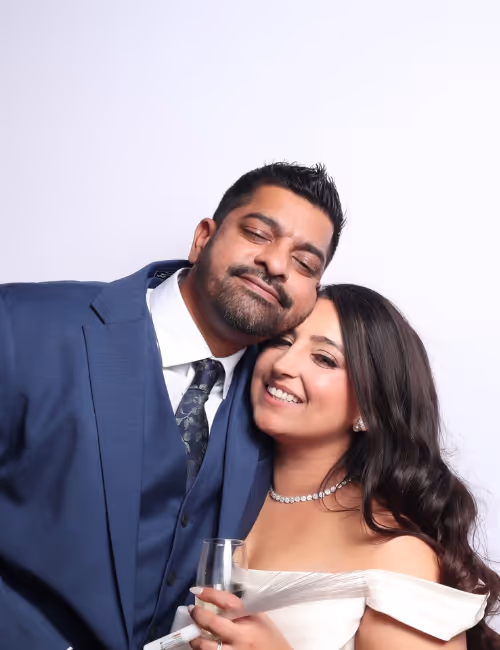 Smiling bride in off-shoulder white dress and groom in blue suit posing closely with eyes closed.