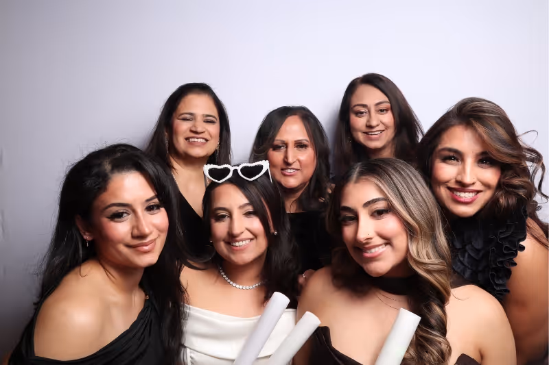 Group of seven smiling women posing together against a plain light background, with two in front holding rolled-up papers and one wearing heart-shaped sunglasses on her head.