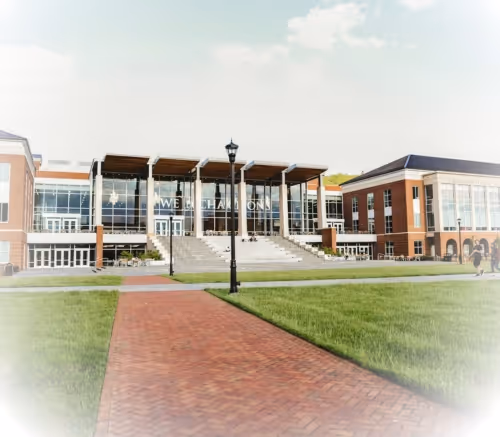 Modern campus building with large glass windows, a central staircase, and a brick pathway leading through a grassy lawn.