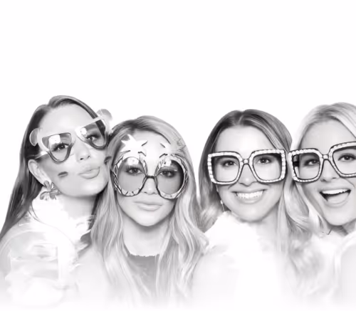 Four women posing in a photo booth with glitter backdrop, wearing Happy New Year headbands and festive outfits