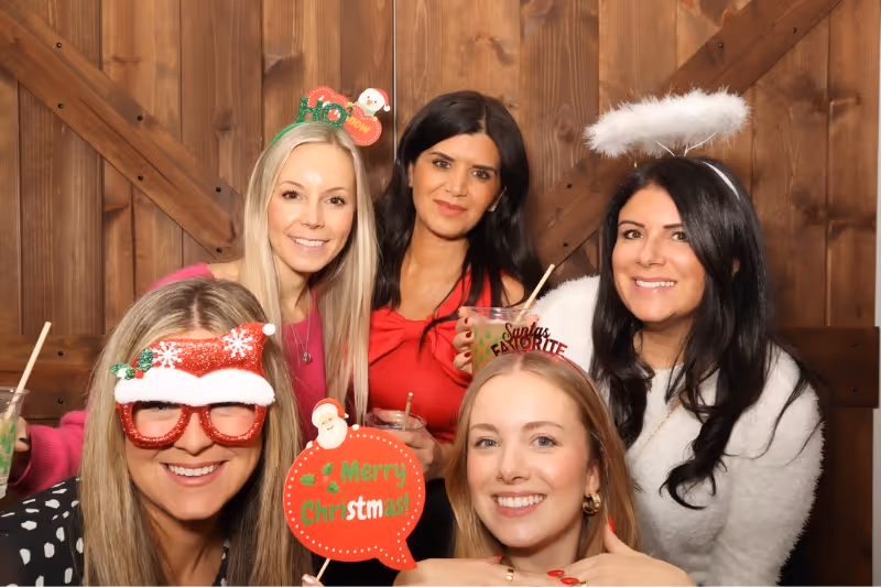 Five women smiling and posing with festive Christmas headbands and props in front of a wooden wall.