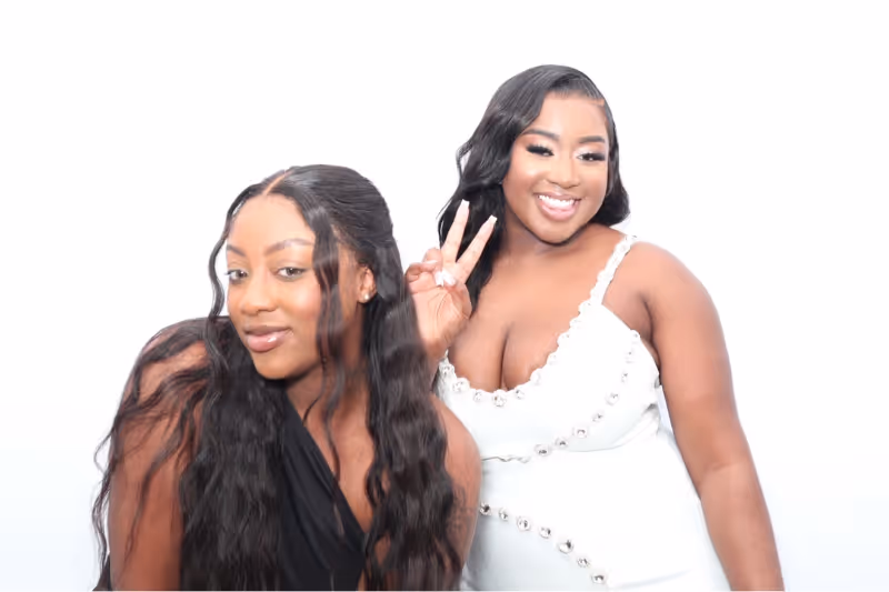 Two women smiling; the one on the right wears a white dress and is making a peace sign with her hand, the one on the left wears a black outfit.