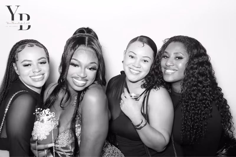 Four smiling women posing closely together, dressed in stylish outfits, with drinks in hand and a logo in the top left corner.