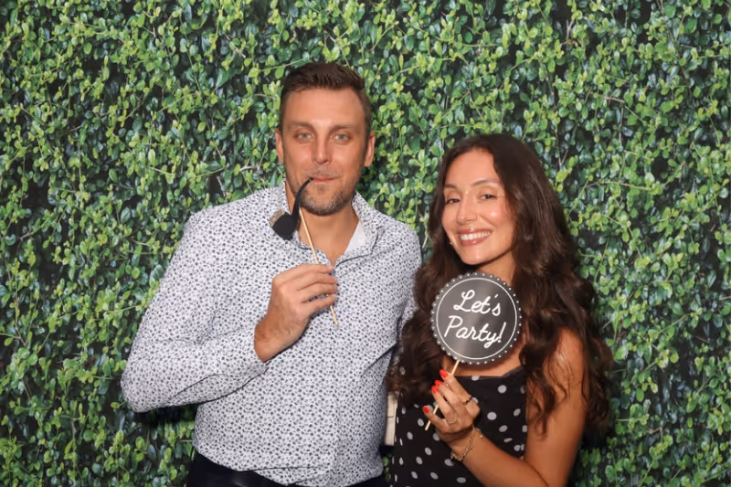 Man and woman smiling in front of leafy green backdrop, holding party photo props including a pipe and a sign that says 'Let's Party!'