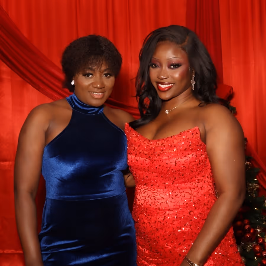 Two women smiling in elegant dresses, one in blue velvet and the other in a red sequined gown, standing against a red curtain backdrop.