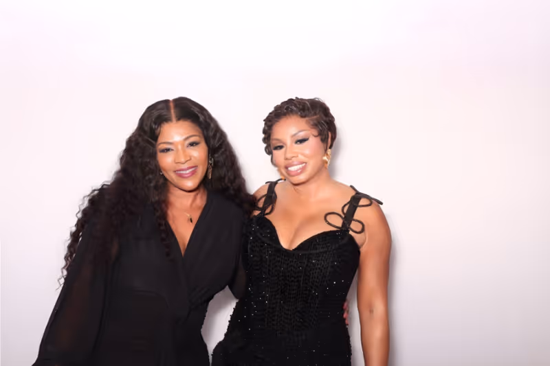 Two smiling women posing together against a plain white background, both dressed in elegant black outfits.