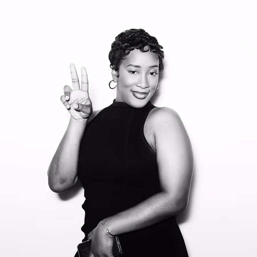 Smiling woman with short curly hair wearing hoop earrings and a sleeveless black top, making a peace sign gesture with her right hand.
