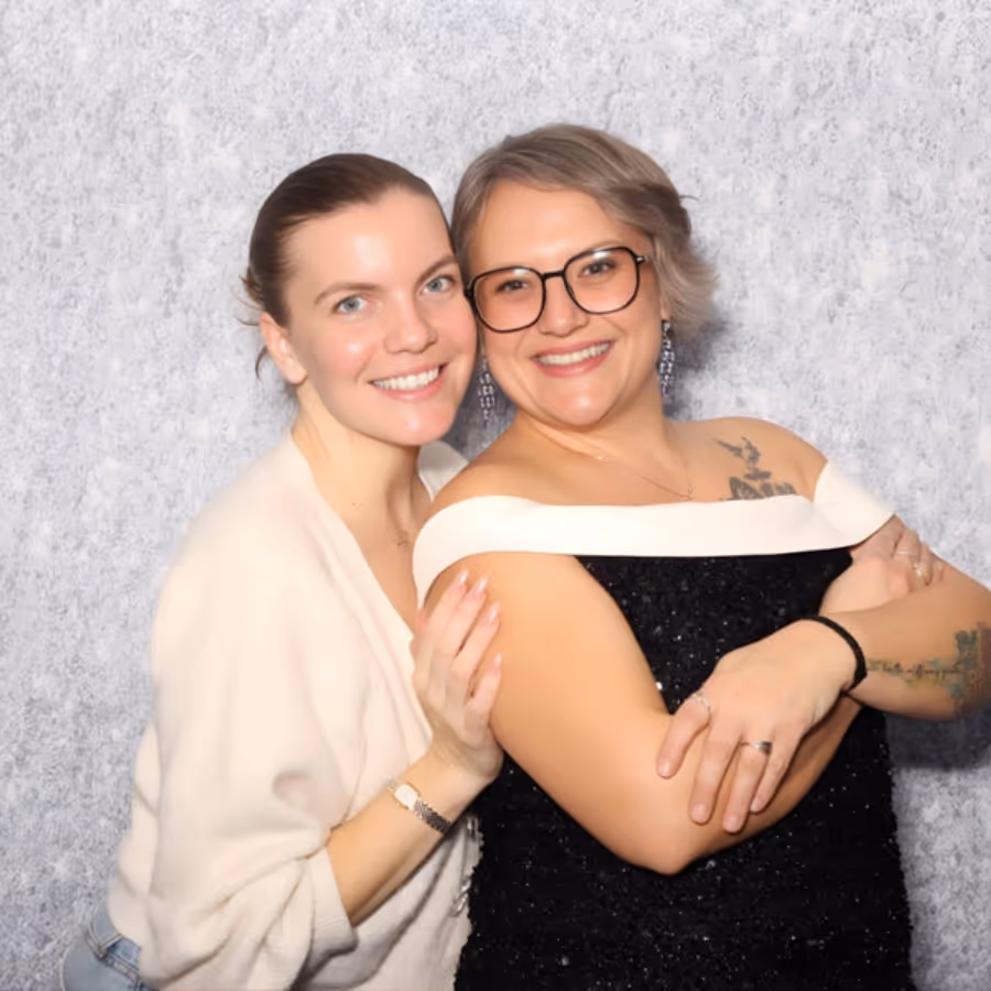 Two women smiling and posing closely together in front of a textured light gray background.