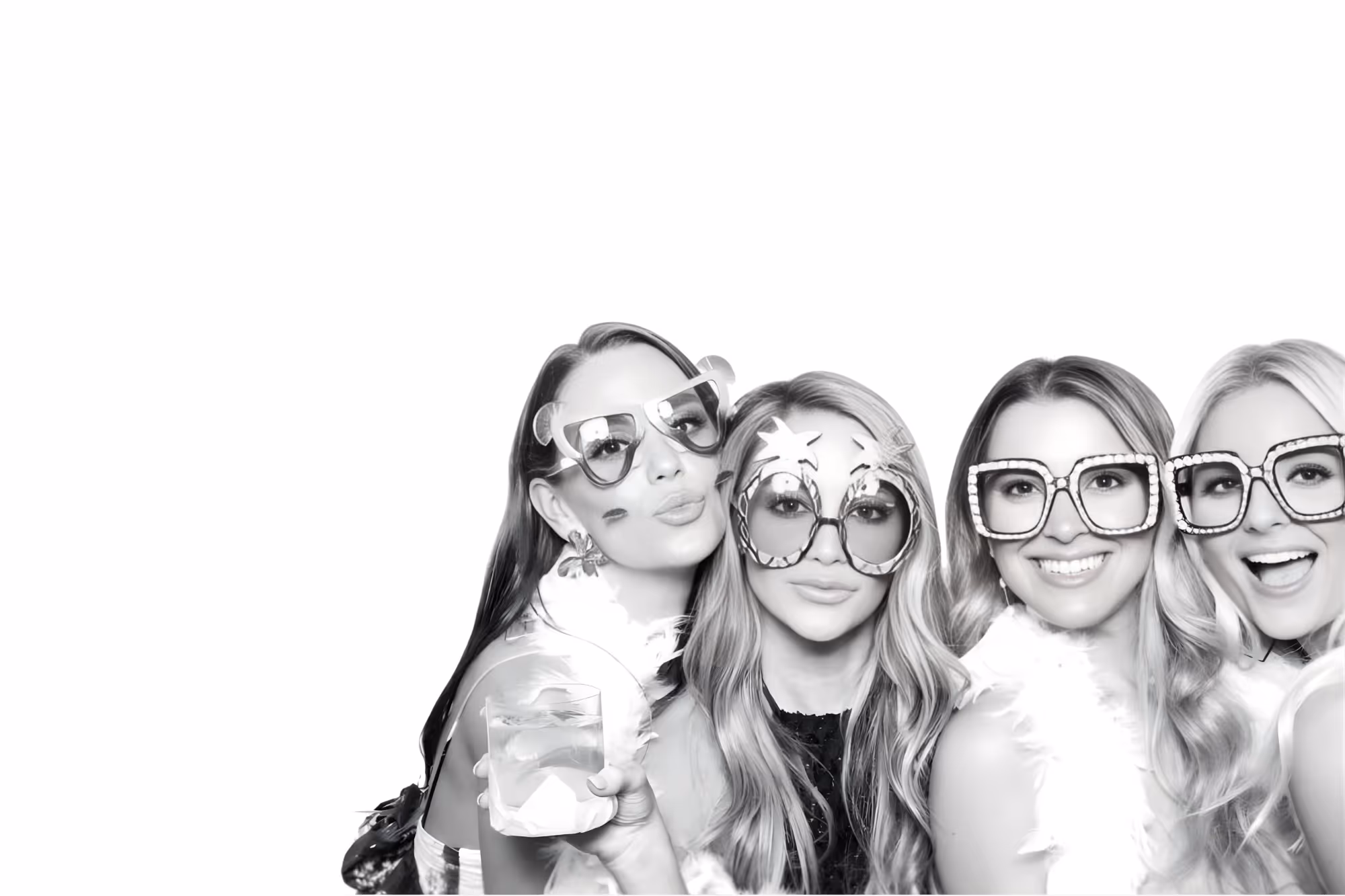 Four women wearing oversized fun sunglasses and feather boas posing together, one holding a drink.