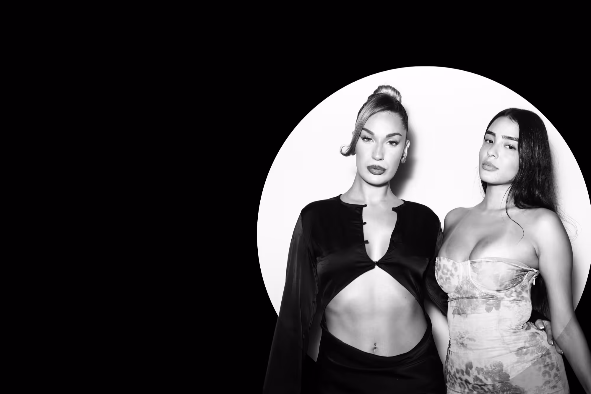 Black and white photo of two women posing confidently against a circular white background, one wearing a black cropped top and the other in a strapless patterned dress.