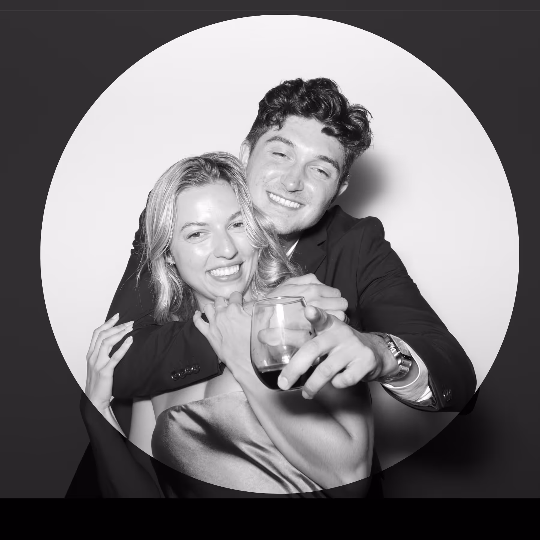 Smiling couple posing closely in a black and white photo, with the man hugging the woman from behind and holding a glass.