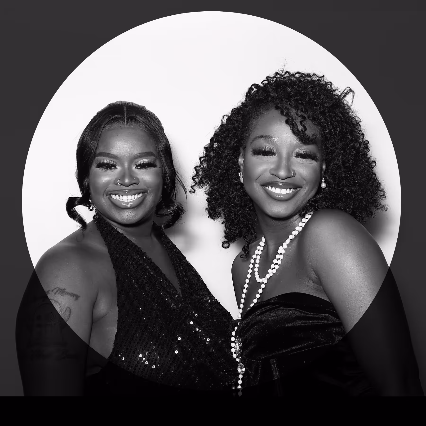 Two smiling women dressed in elegant black attire, one wearing a pearl necklace and the other with a nose ring and visible arm tattoo.