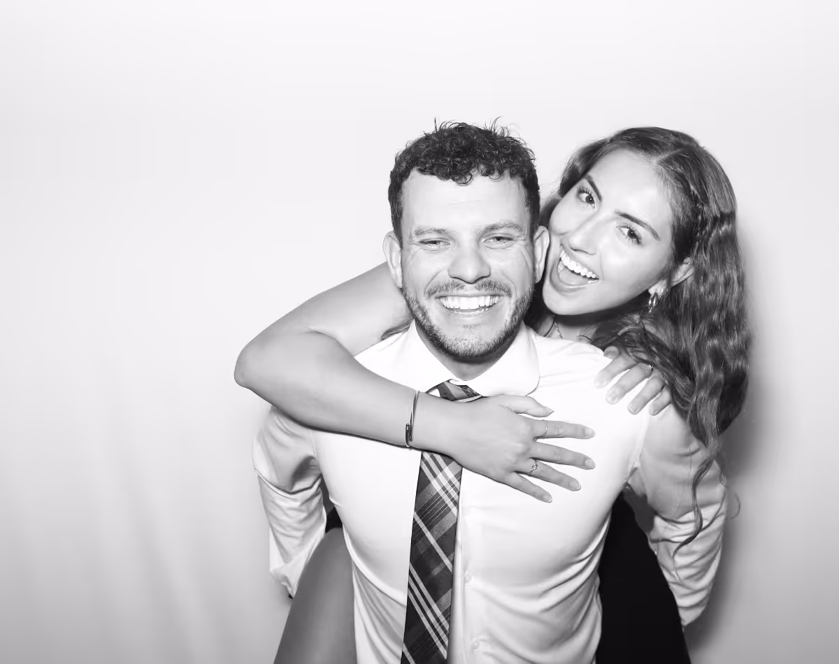 Smiling woman with long hair hugging a man in a dress shirt and plaid tie from behind against a plain background.