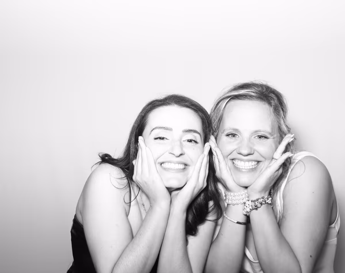 Two smiling women resting their faces in their hands, posing closely together against a plain background.