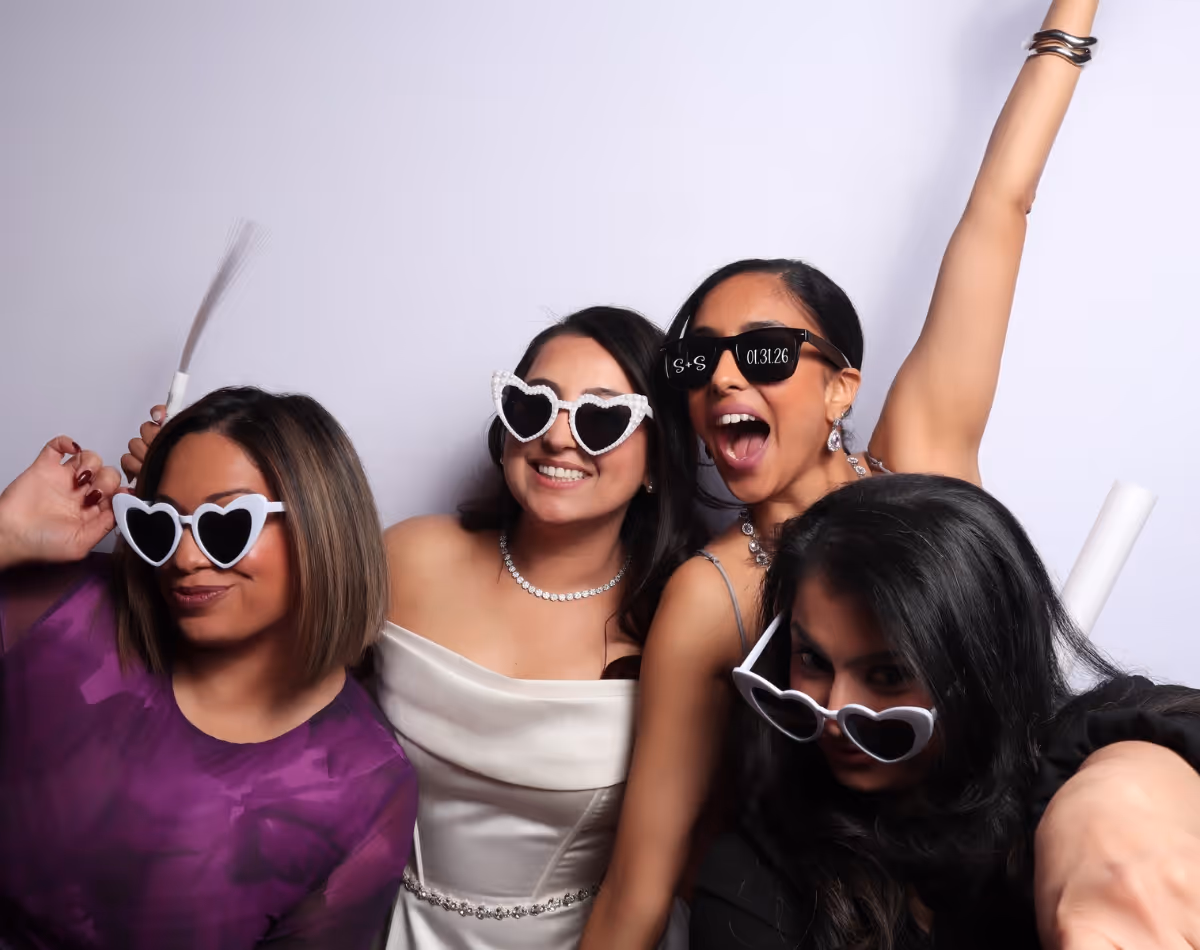 Four women wearing heart-shaped sunglasses posing happily against a plain light background.