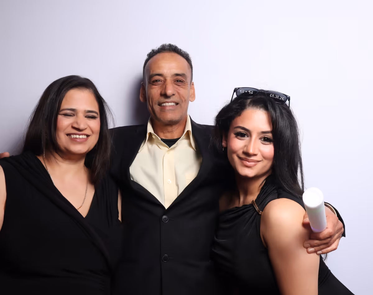 Three people posing closely together smiling in front of a plain light background; two women in black dresses and a man in a black suit with a cream shirt.