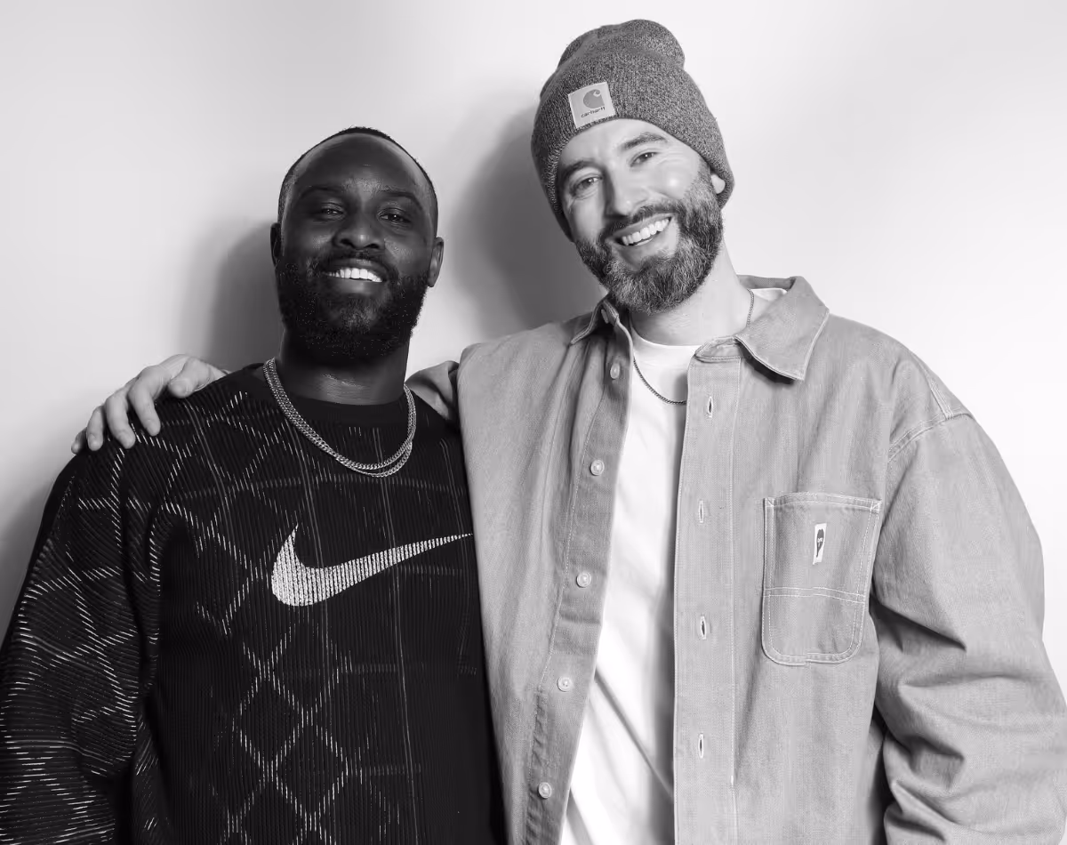 Two men smiling and posing together, one wearing a beanie and casual jacket, the other in a patterned Nike sweatshirt.
