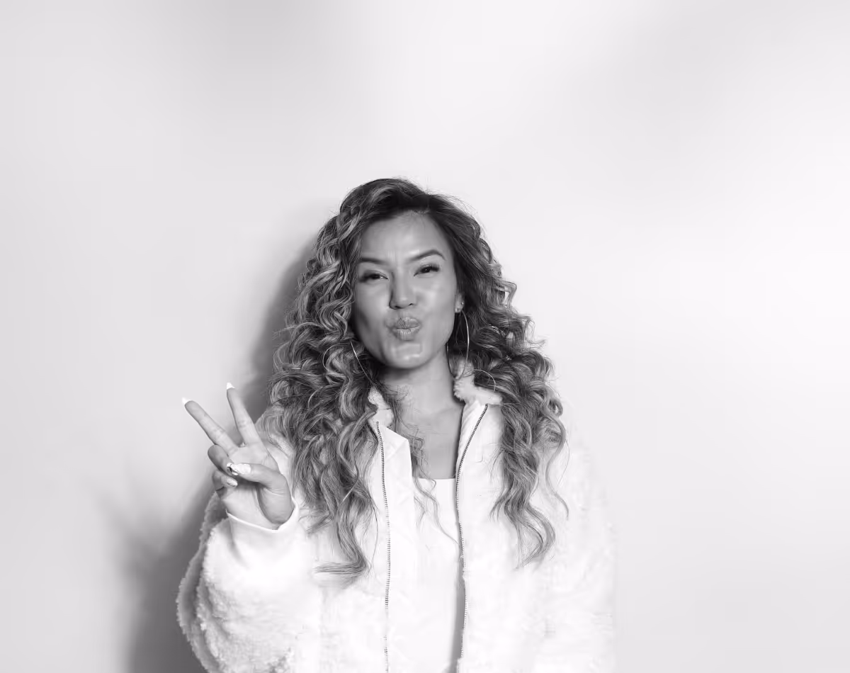 Woman with long curly hair wearing a fluffy jacket making a peace sign and puckering lips in a playful pose.
