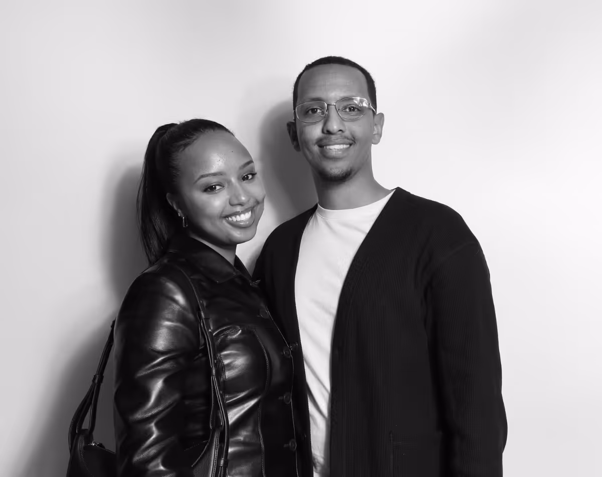 Smiling couple standing close together against a plain background, the woman wearing a leather jacket and the man wearing glasses and a cardigan.
