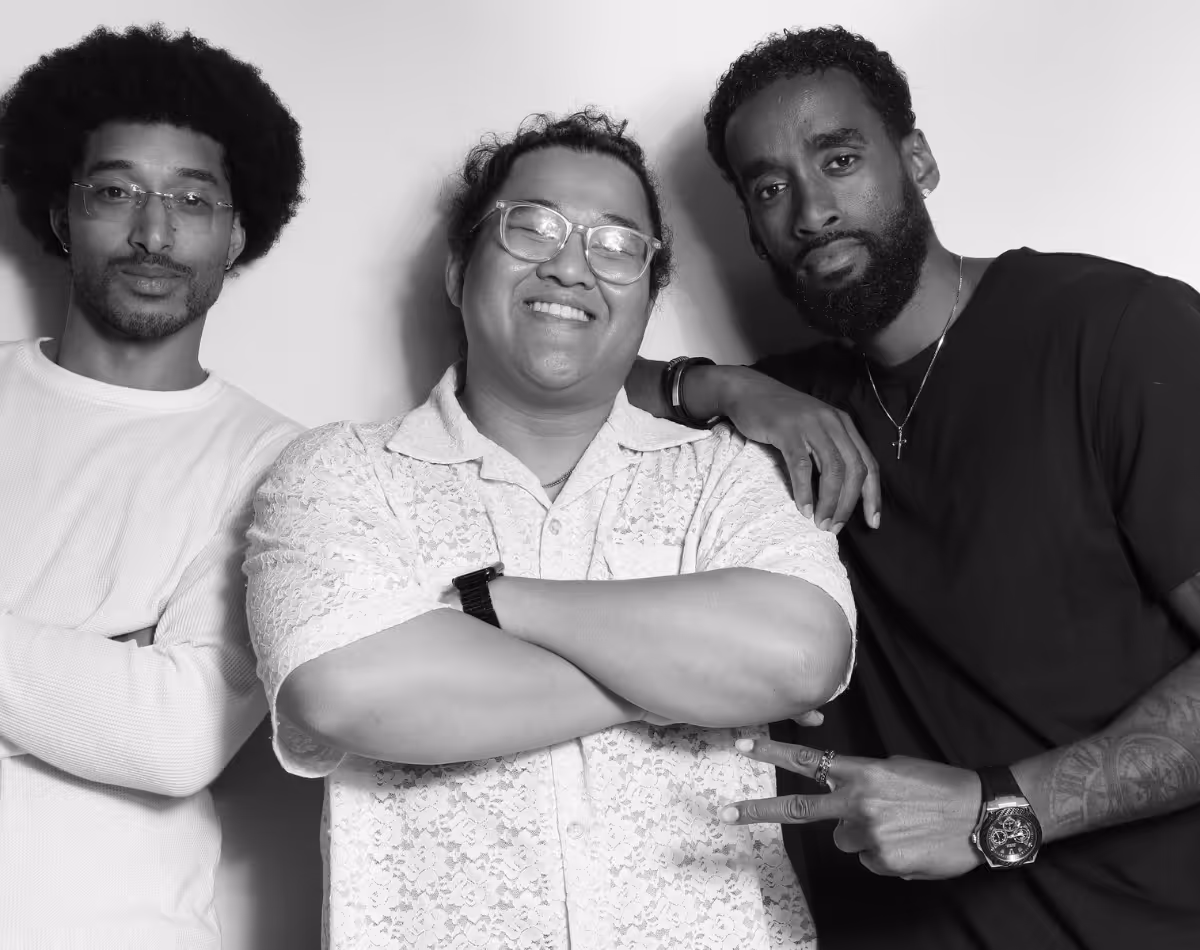Three men posing together, with one in the center smiling with crossed arms, the man on the right making a peace sign and wearing a watch, and the man on the left with glasses and arms crossed.