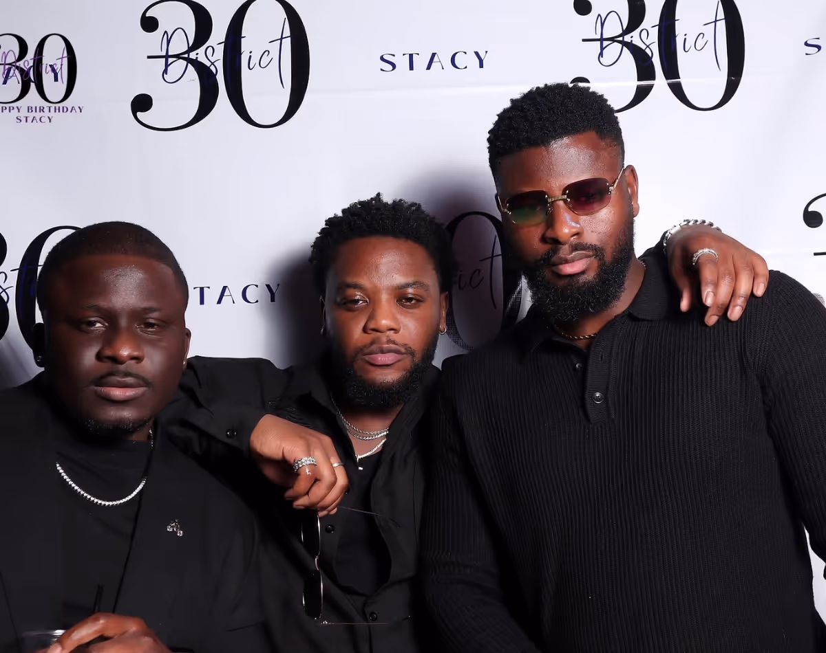 Three men dressed in black posing together in front of a backdrop with '30 Bistrict Happy Birthday Stacy' text.