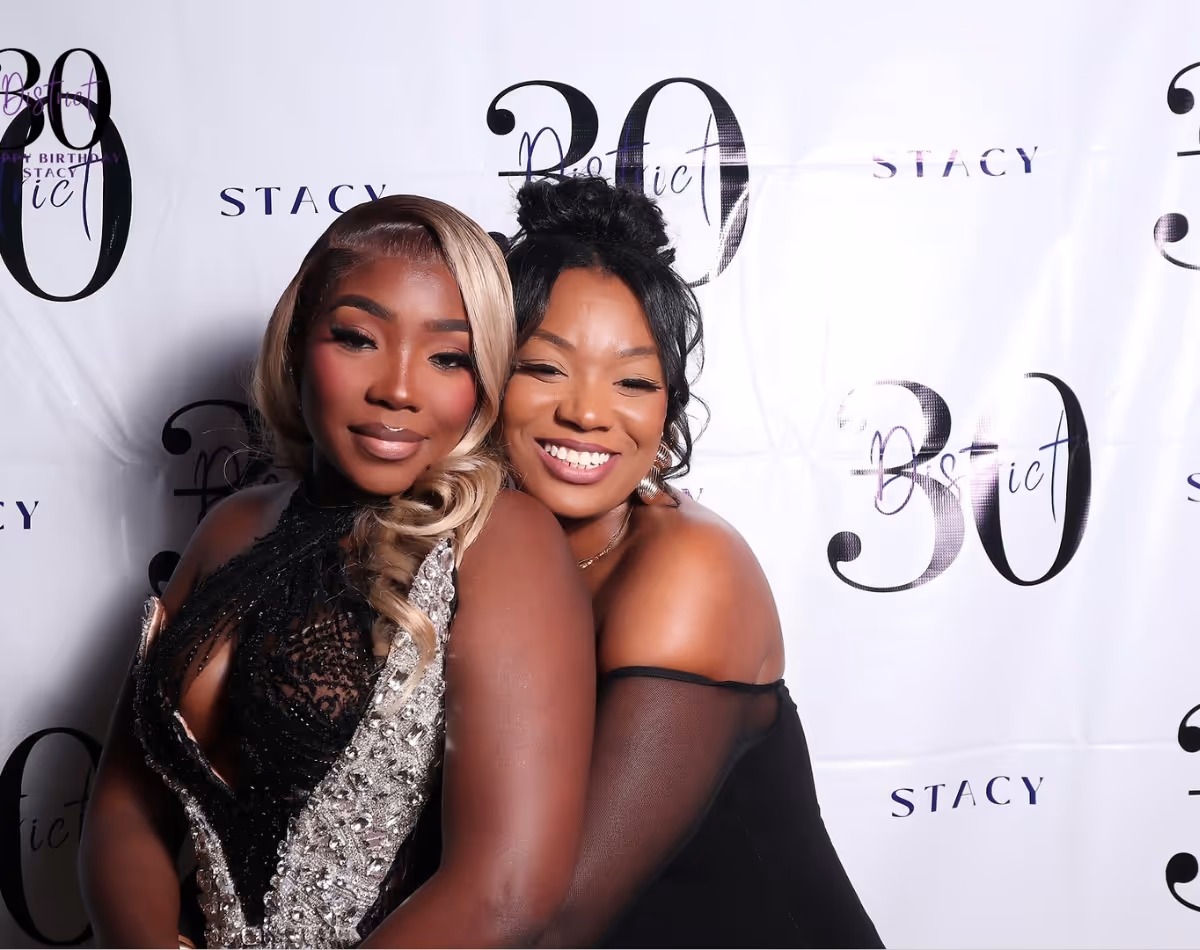 Two women smiling and embracing at a celebration with a backdrop reading '30 Stacy'.