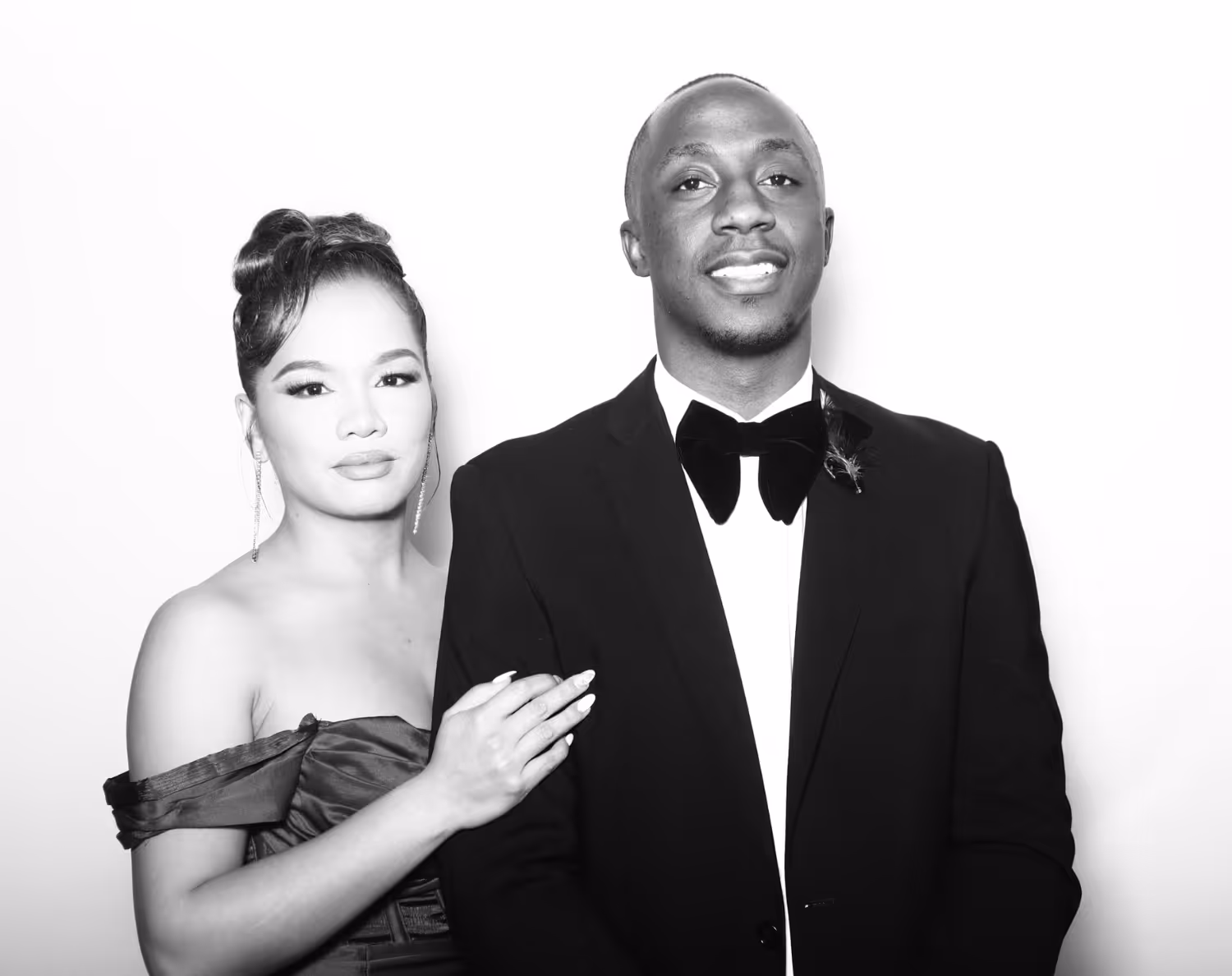 Elegant couple in formal attire posing; woman in off-shoulder dress with styled hair, man in tuxedo with bow tie.