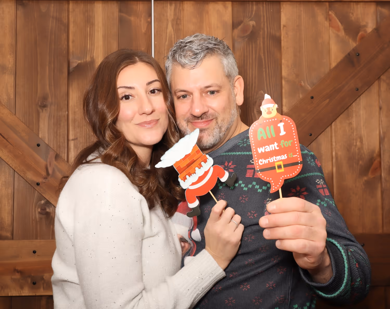 Smiling couple wearing holiday sweaters holding Christmas-themed photo props in front of a wooden backdrop.