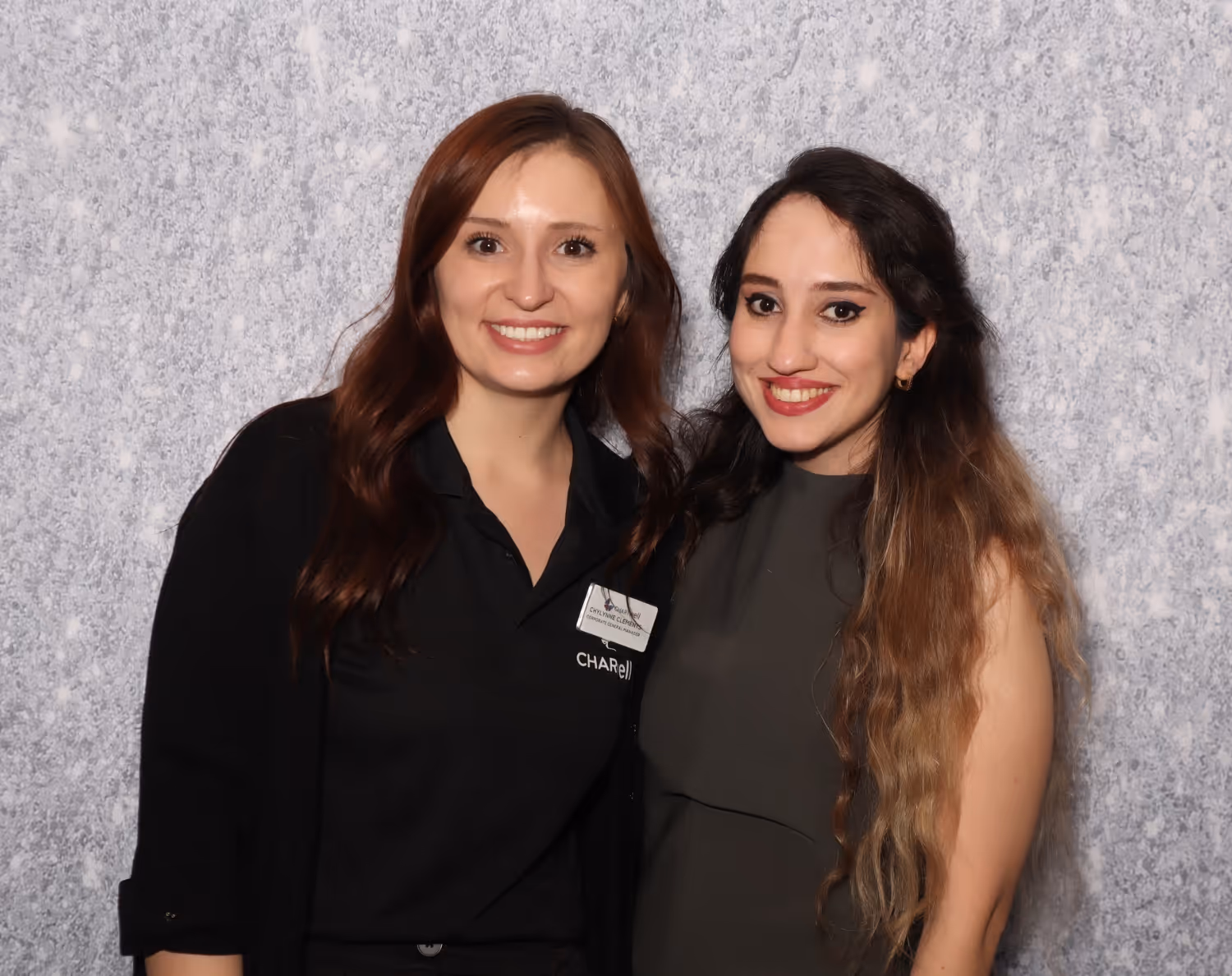 Two smiling women standing close together against a light gray textured background, one wearing a black collared shirt and the other in a dark sleeveless top.