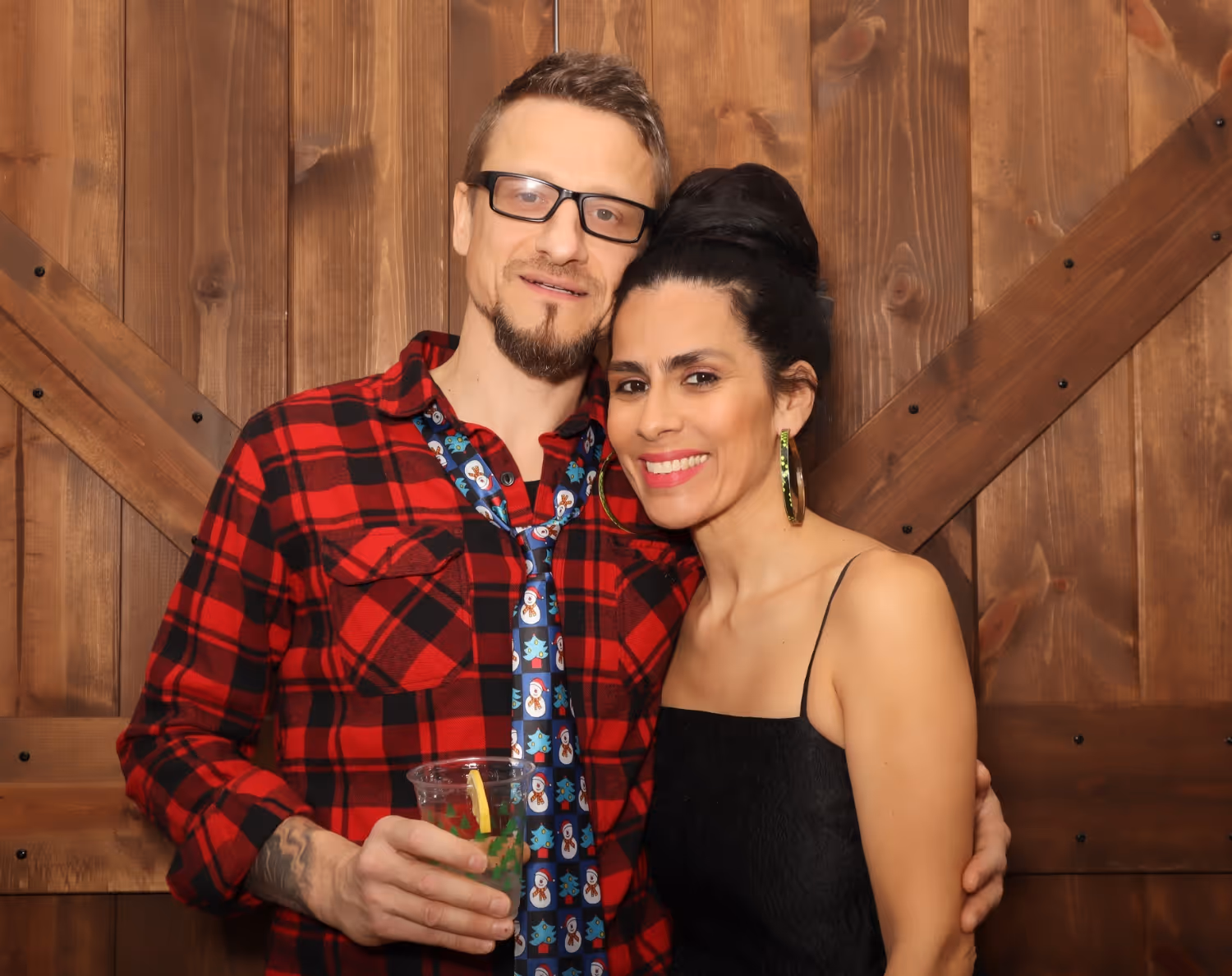 Smiling couple posing indoors in front of wooden wall; man in red plaid shirt and festive tie holding a drink, woman in black dress with large hoop earrings.