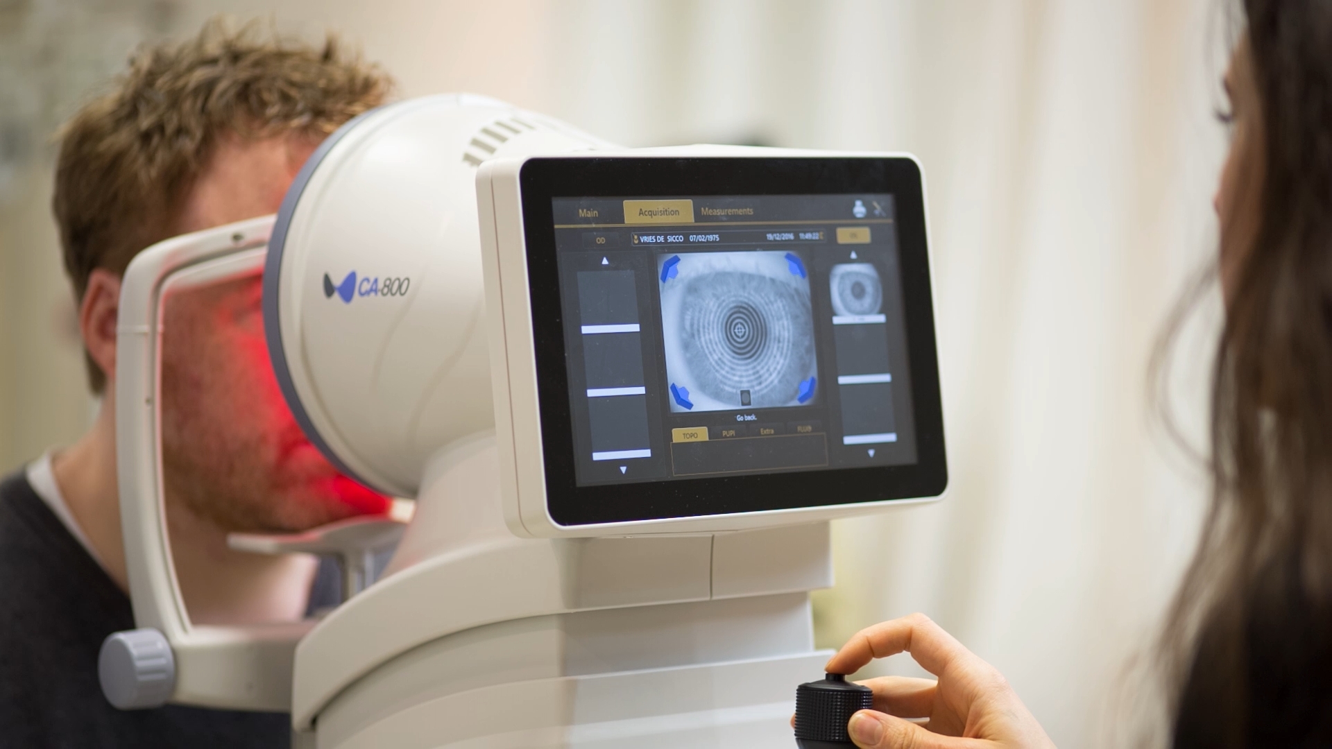Patient undergoing an eye examination using a CA800 corneal topographer with a close-up of the device display showing an eye scan.