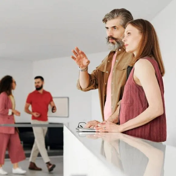deux personnes, une homma et une femme, en premier plan qui regardent au loin avec un plan de travail pour s'appuyer. Deux autres personnes en secon plan avancent dans l'espace cuisine en discutant