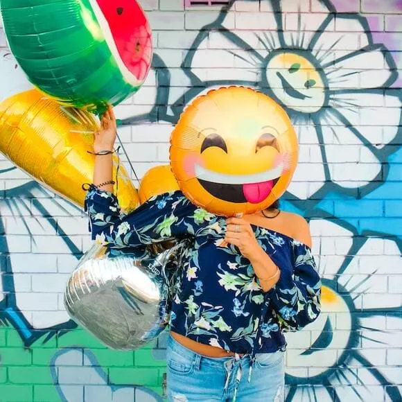 jeune fille avec ballon happy face devant son visage et un tenant un autre ballon en forme de pasteque dans son autre main, se trouvant debout dans une rue avec un mur, derrière elle, avec un beau tag de fleurs