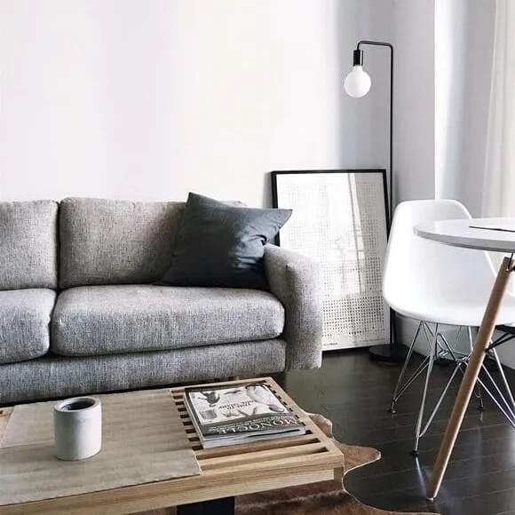 appartement meublé avec un canapé gris, une table basse en bois avec une tasse et un magazine, en arrière plan un tableau à même le sol et une lampe. Sur la droite un table avec une chaise moderne