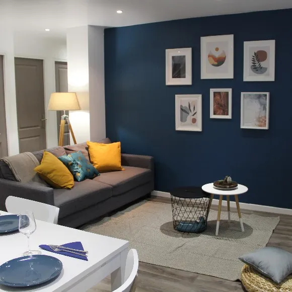 salon avec mur bleu et cadres de décoration, un canapé gris avec des coussins jaune et vert, une table blanche en premier plan avec des assiettes bleues et des couvert