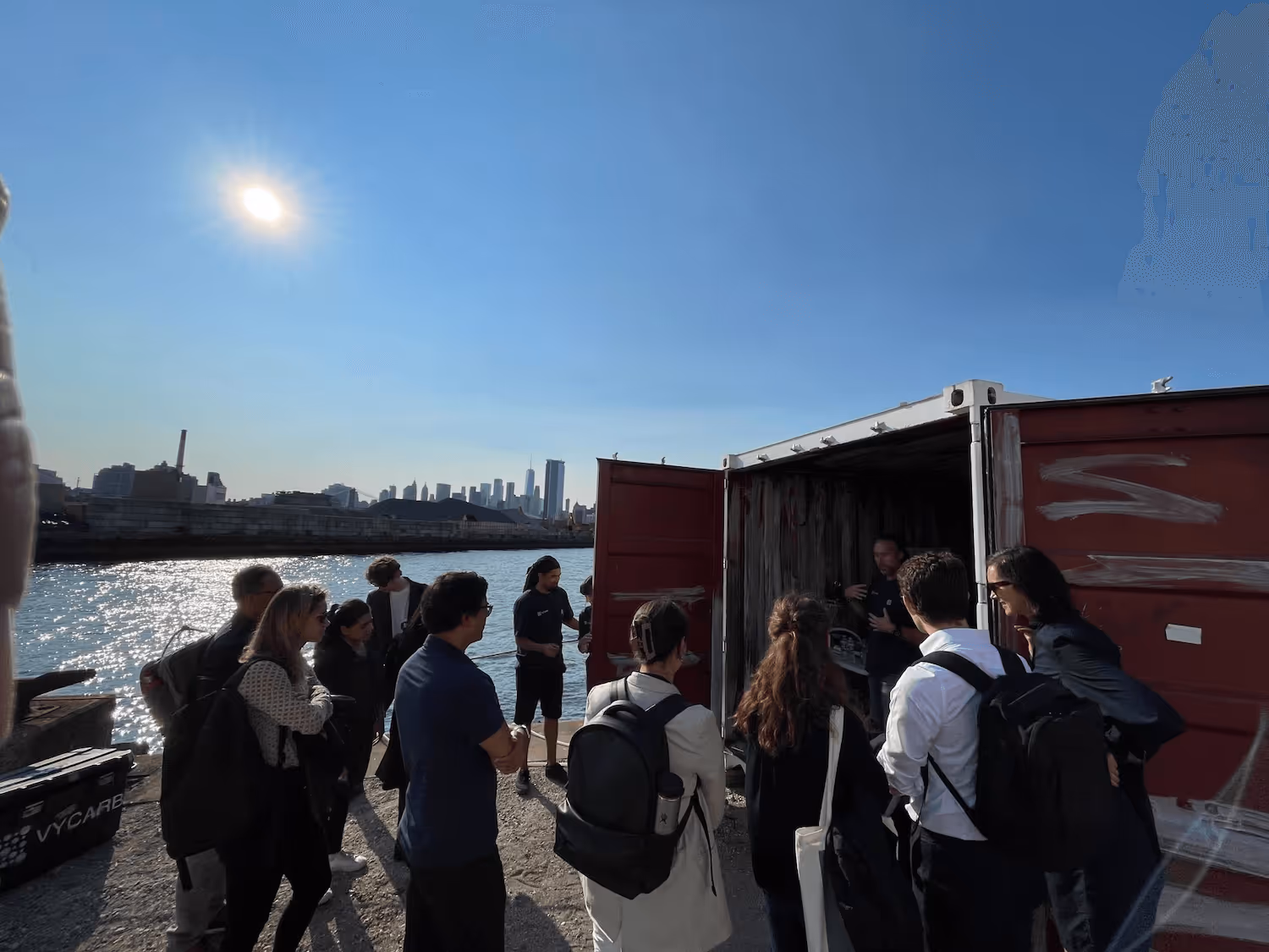 Group of people gathered outdoors near a waterfront, listening to a man speaking inside an open red shipping container with a city skyline in the background under a clear sky.