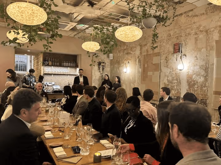Group of people seated around a long wooden table in a warmly lit room with hanging plants and round ceiling lights, listening to a person speaking near the back wall.