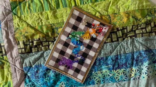 Artwork description: Overhead view of Mend To Reconstruct, a small quilt made out of beads, unframed, laying on a colourful, multi patterned fabric quilt. The art piece is a beaded version of a gingham quilt in high contrast of transparent white, metallic brown, and opaque black. A rainbow gradient of patches of “mending” are beaded across the piece. The colourful “mending” beads are primarily small with some larger round and unique shaped beads included.