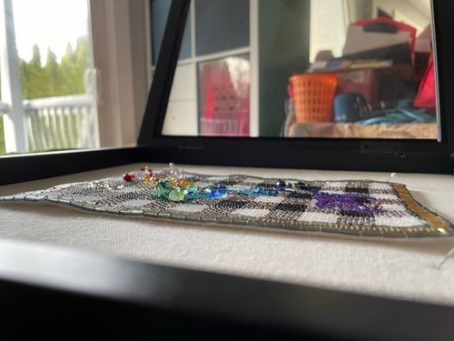 Artwork description 4: An angled close up of the front of the art piece laying, in the black framed shadow box. The raised textures of the beads, especially the colourful, larger shaped beads in different shapes are visible. The background of the room is slightly more in focus with white shelving and a pile of things visible.