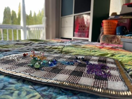Artwork description 5: A different angled close up of the front of the art piece laying, unframed, on a colourful, multi patterned fabric quilt on a bed. The raised textures of the beads, especially the colourful, larger shaped beads in different shapes are visible. The purple side of the colourful beads in the piece are on the right of the image, the red and white beaded sides on the left. In the background, the room with white shelving and a pile of things are more visible.