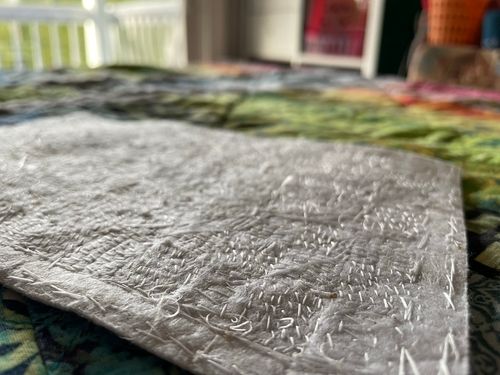 Artwork description 6: A detailed, unframed close up image of the back of the beaded quilt at an angle. Stitches, knot ties and several tiny burn marks where thread was melted to strengthen fraying point are visible on the white backing batting.
The piece is laying flat on the colourful fabric quilt. A room out of focus is in the background.