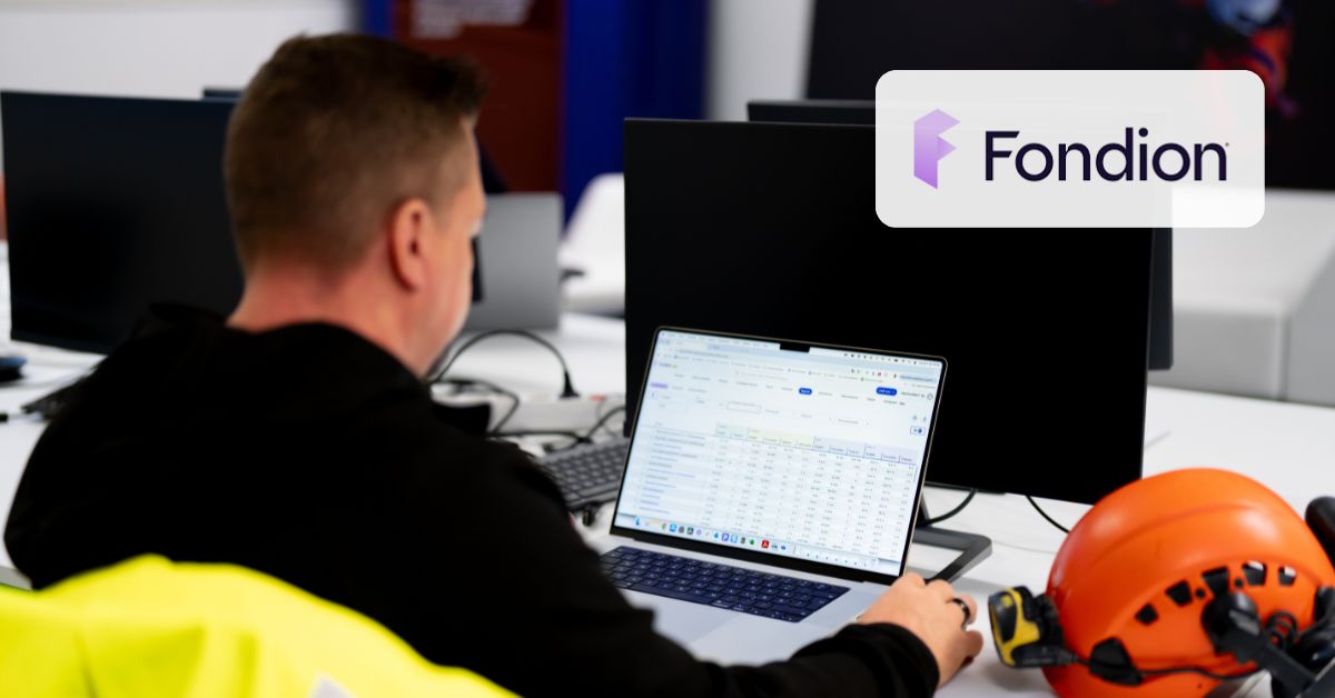 Henkilö istuu koneen äärellä ja Fondion ERP-ohjelmisto on auki hänen edessään. Pöydällä työmaakypäräja ja kuvassa myös huomiotakki.