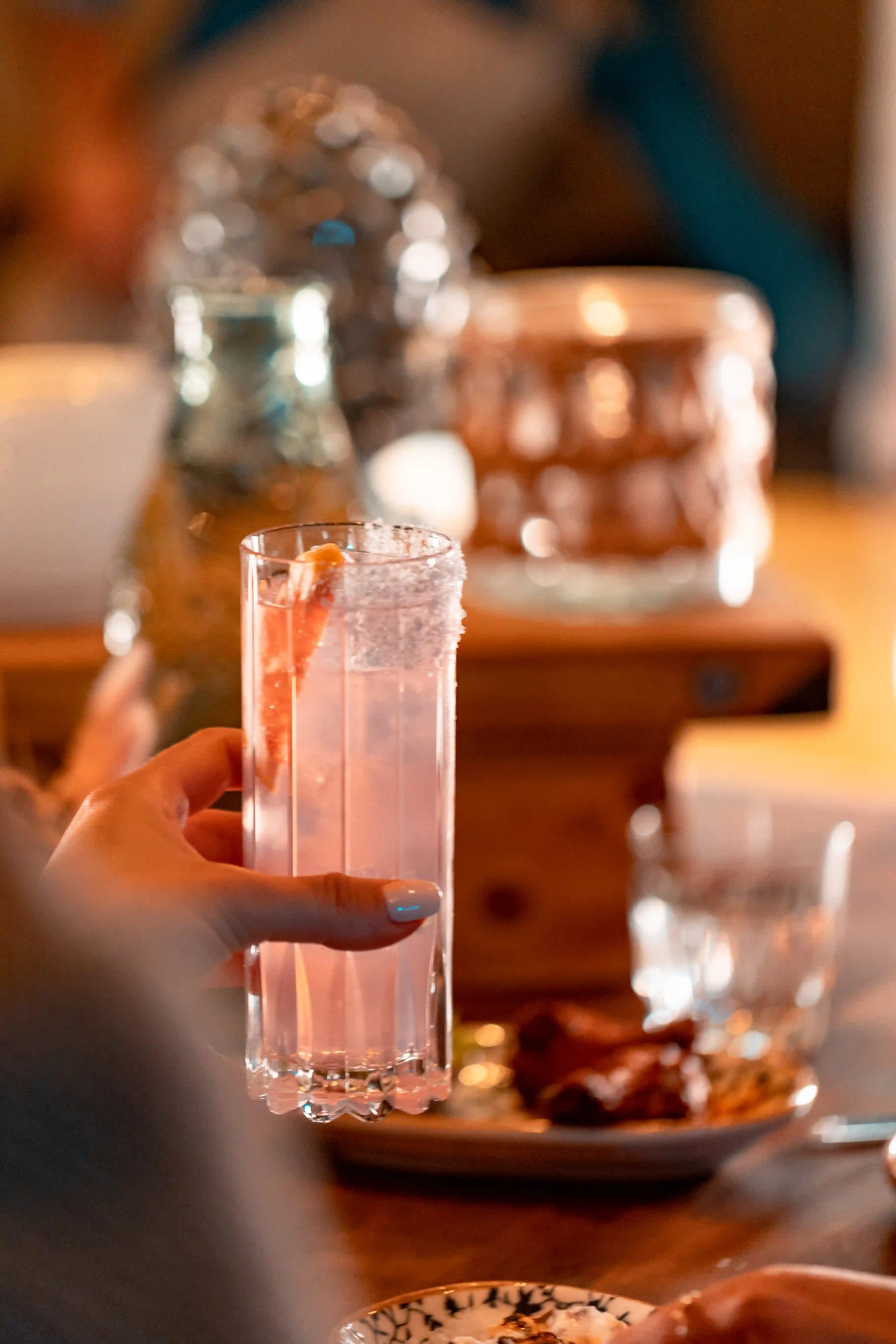 Hand som håller ett högt glas med en rosa cocktail och en citrusskiva, med en oskarp bakgrund av ett bord och dekorativa föremål